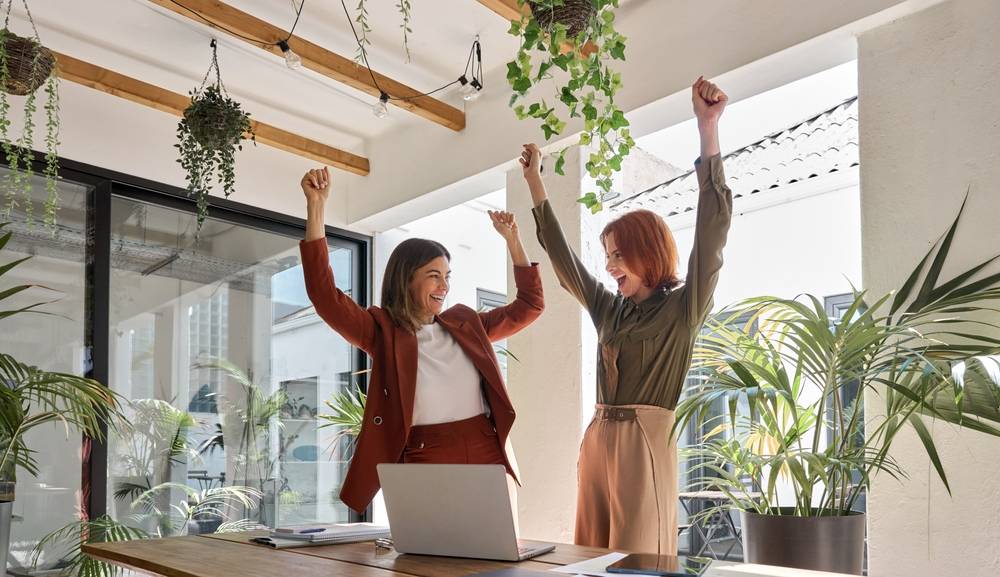 Dve žene u kancelariji, stoje, digle su ruke u vazduh i raduju se nekom rezultatu.