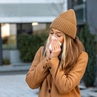 Žena u kaputu, nosi kapu i briše nos maramicom.