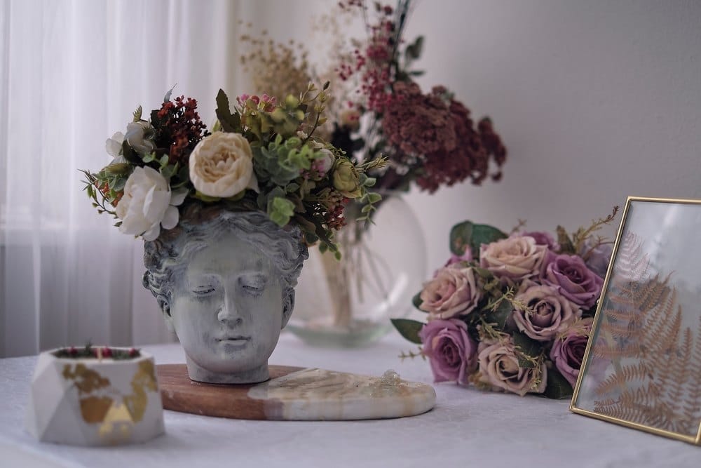 Vaza koja je u obliku glave Afrodite, iz nje viri veliki buket raznog cveća, pored na stolu stoji još jedan buket ruža.