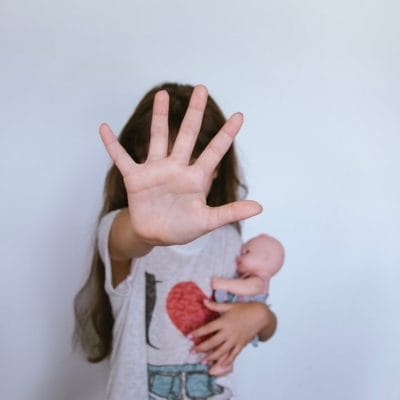 Devojčica drži lutku, lice joj s ene vidi, jer joj je ruka ispred lica, kao da pokazuje STOP.