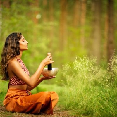 Žena koja sedi u šumi u sariju. U rukama drži specijalnu staklenu činiju i palicu kojom dodirom činije proizvodi zvuk. Vrlo je opuštena.