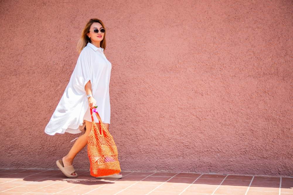 Devojka u beloj haljini-kosulji. Nosi narandžastu torbu, ima naočare za sunce.