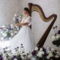 Sofija Sibinović u dugoj beloj haljini od tila, stoji pored harfe, okružena belim i ljubičastim cvećem,