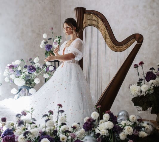 Sofija Sibinović u dugoj beloj haljini od tila, stoji pored harfe, okružena belim i ljubičastim cvećem,