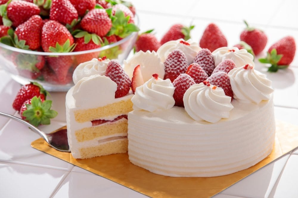 Torta sa jagodama, ukrašena šlagom. Jedno parče je izvađeno i vidi se presek torte, kore, fil i jagode.