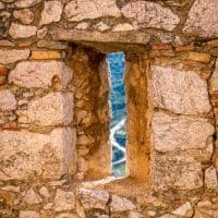 Mali prozor u zidu tvrđave, kroz koji se nazire krivudavi put.