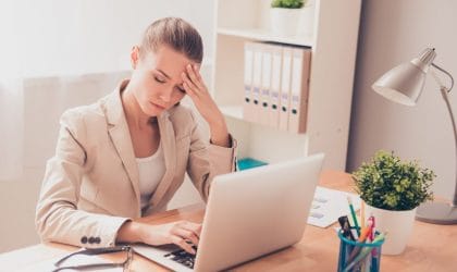 Premorena žena u četrdesetim sedi u kancelariji za stolom. Umorna je, drži se za glavu.