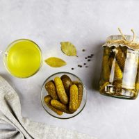 Tegla sa kiselim krastavcima, kiseli krastavci u činijici, lovorov list, a pored je čaša sa sokom od kiselih krastavaca.