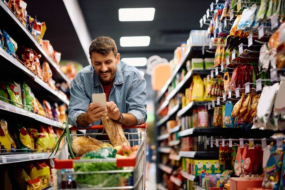Čovek u supermarketu naslonjen na kolica gleda u spisak za kupovinu na svom telefonu.