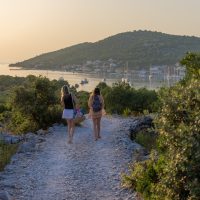 Žene koje hodaju kamenom stazom, u daljini se vidi gradić na moru.