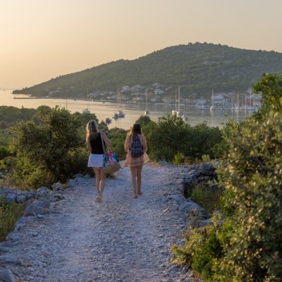 Žene koje hodaju kamenom stazom, u daljini se vidi gradić na moru.