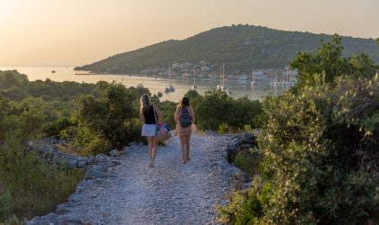 Žene koje hodaju kamenom stazom, u daljini se vidi gradić na moru.