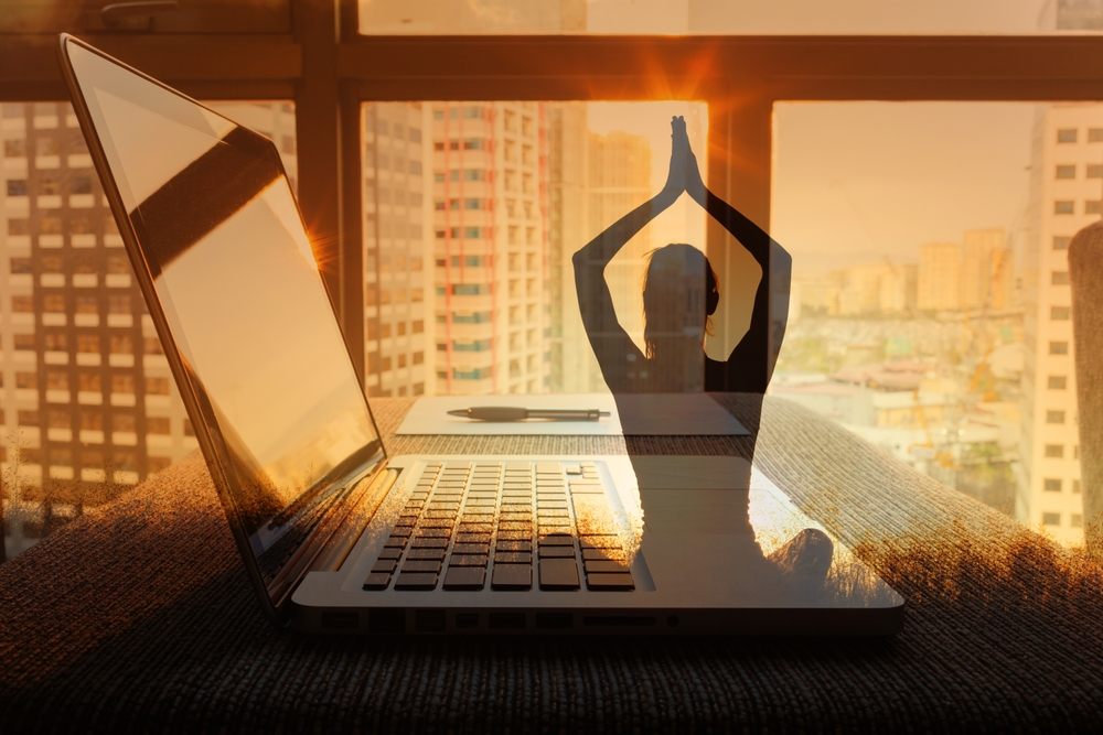 Fotografija ilustracija, lap top na stolu, kroz prozor se vidi grad. Preko fotografije je ilustracija žene koja radi jogu.