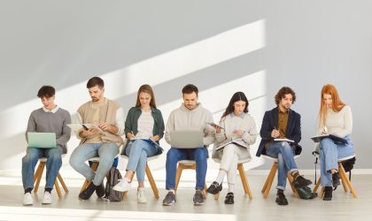 Grupa ljudi, muškarci i žene, sede na stolicama, popunjavaju prijave za posao.