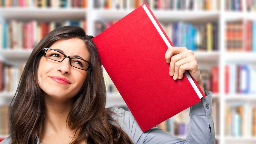 Žena u biblioteci naslonila je knjigu na glavu i kao da promišlja i da je došla do nekog zaključka.