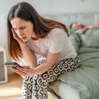 Zabrinuta žena sedi na ivici bračnog kreveta u kome spava njen partner. Ona zabrinuto gleda u telefon.