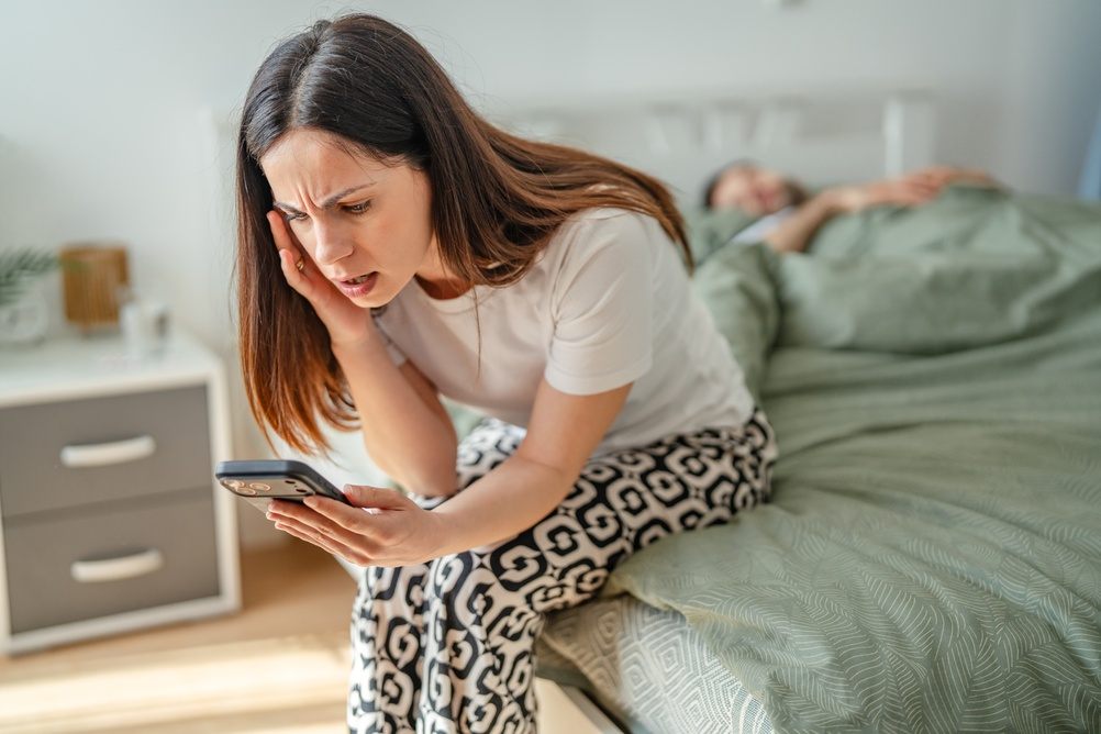 Zabrinuta žena sedi na ivici bračnog kreveta u kome spava njen partner. Ona zabrinuto gleda u telefon.