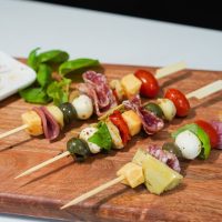 Antipasto ražnjići. Na drvene štapiće nataknut je čeri paradajz, krastavac, sir, masline i dr. Postavljeni su na dasci.