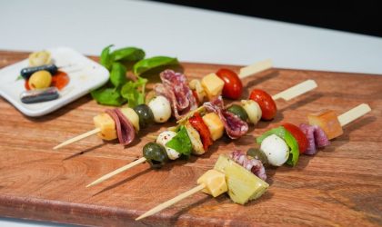 Antipasto ražnjići. Na drvene štapiće nataknut je čeri paradajz, krastavac, sir, masline i dr. Postavljeni su na dasci.