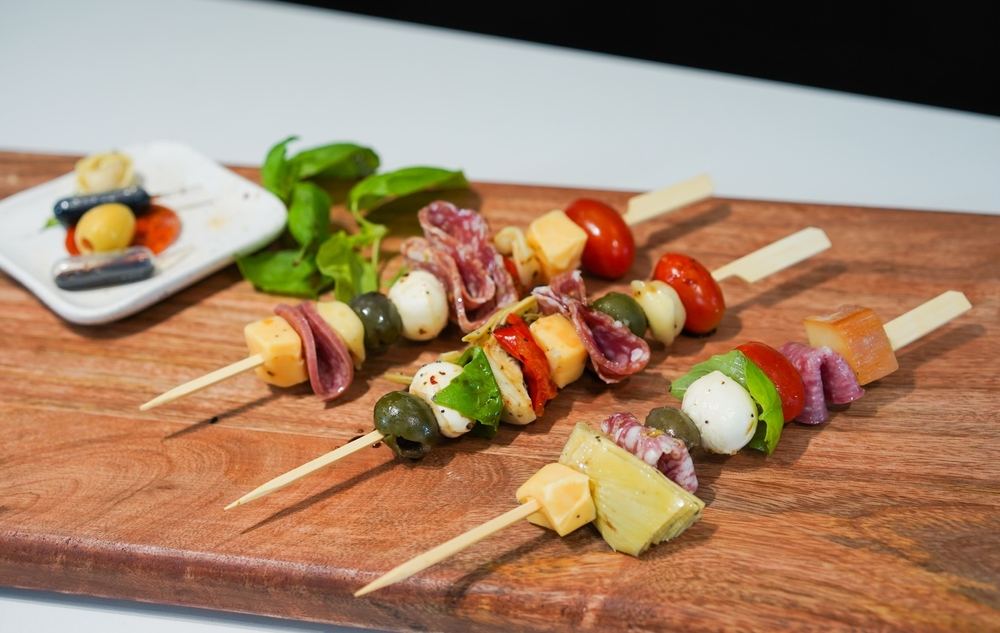 Antipasto ražnjići. Na drvene štapiće nataknut je čeri paradajz, krastavac, sir, masline i dr. Postavljeni su na dasci.