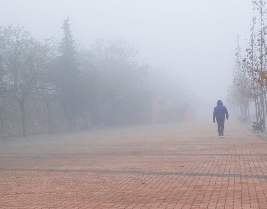 Čovek koji se vidi s leđa, nosi jaknu sa kapuljačom. Hoda pločnikom. Okolo i u daljini je magla.