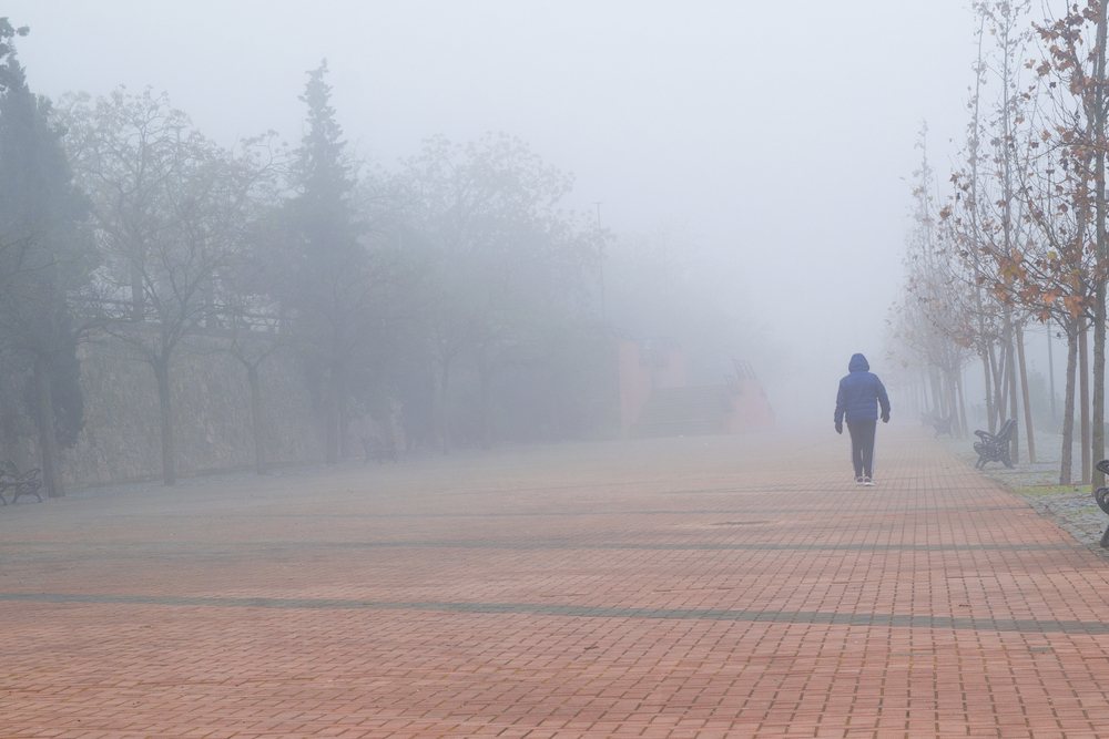 Čovek koji se vidi s leđa, nosi jaknu sa kapuljačom. Hoda pločnikom. Okolo i u daljini je magla.