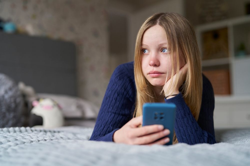 Devojčica leži na krevetu, u ruci drži mobilni telefon i tužno je zagledana u daljinu.