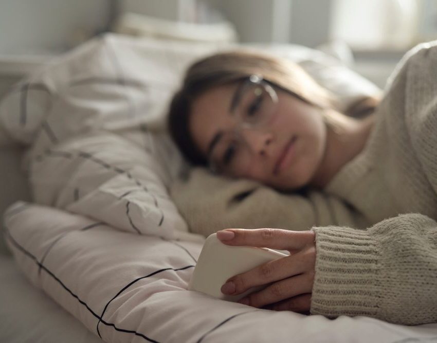 Devojka sa naočarima leži u krevetu i gleda u mobilni telefon.