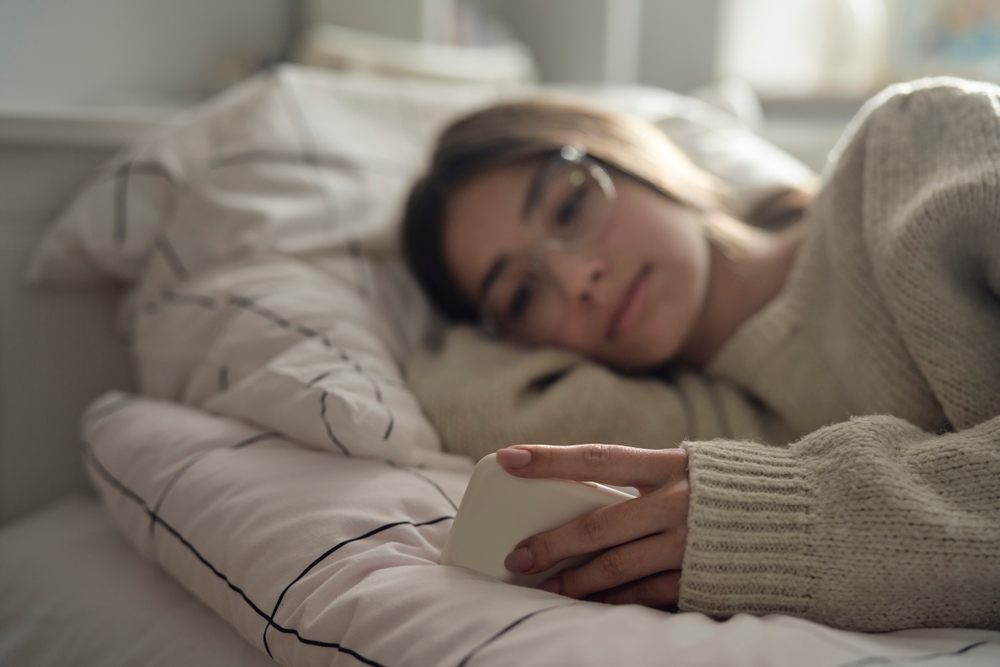 Devojka sa naočarima leži u krevetu i gleda u mobilni telefon.