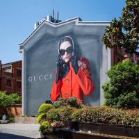 Gucci reklama-bilbord na zidu kuće. Žena u jarko crvenom kaputu, nosi velike crne naočare za sunce.