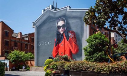 Gucci reklama-bilbord na zidu kuće. Žena u jarko crvenom kaputu, nosi velike crne naočare za sunce.