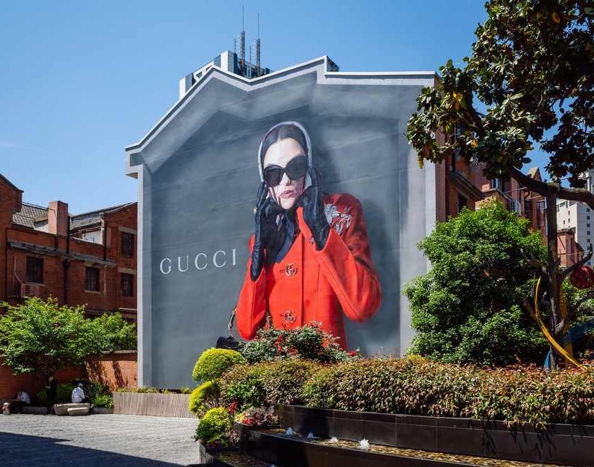 Gucci reklama-bilbord na zidu kuće. Žena u jarko crvenom kaputu, nosi velike crne naočare za sunce.