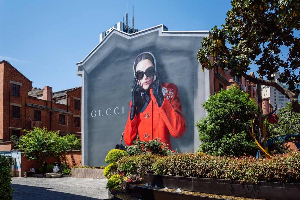 Gucci reklama-bilbord na zidu kuće. Žena u jarko crvenom kaputu, nosi velike crne naočare za sunce.