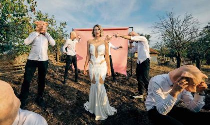 Ana Stanić u prirodi, oko nje su muškarci u crnim pantalonama, belim košuljama, nose gumene maske sa likom praseta.