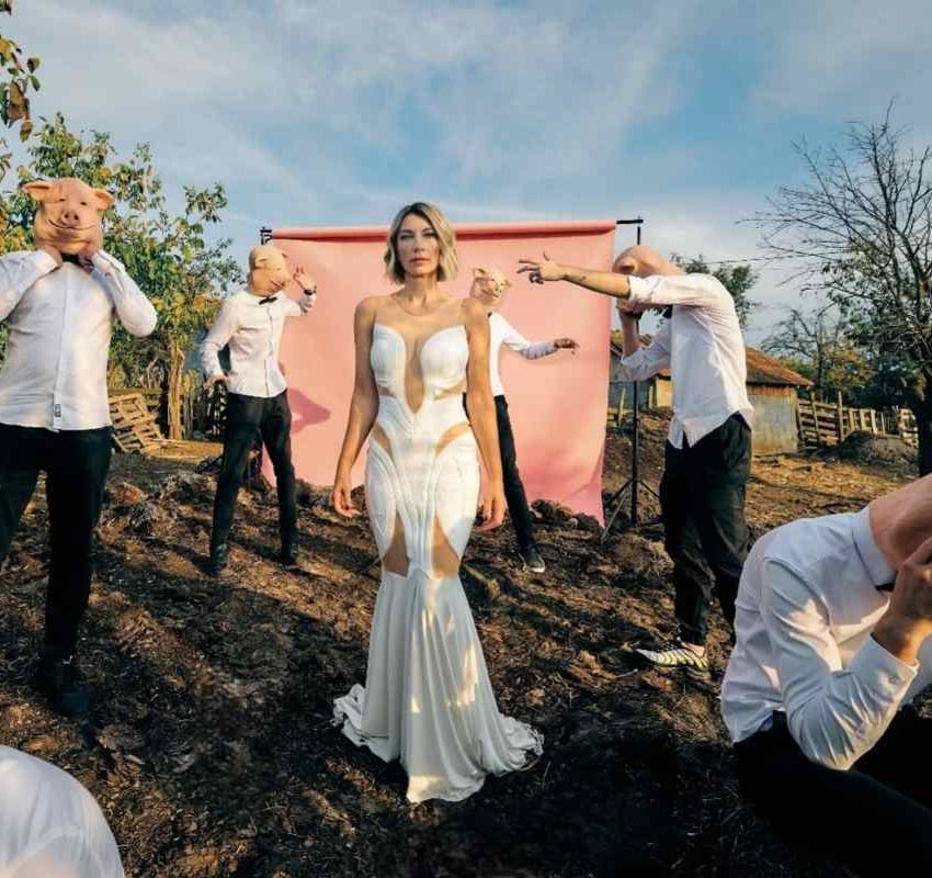 Ana Stanić u prirodi, oko nje su muškarci u crnim pantalonama, belim košuljama, nose gumene maske sa likom praseta.