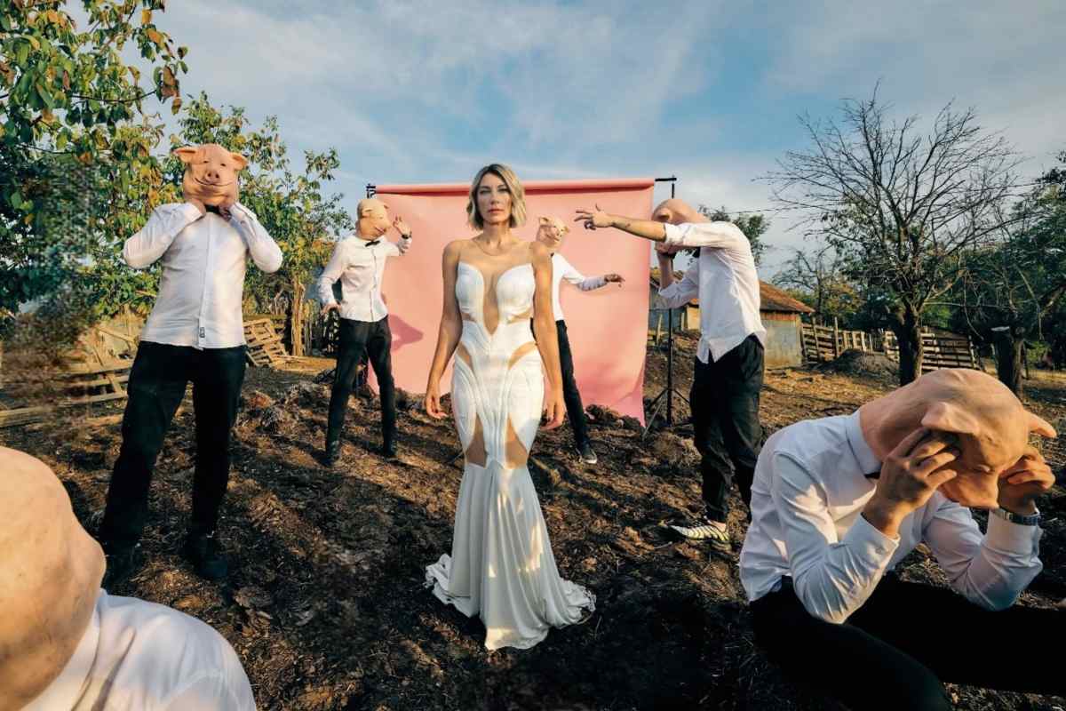 Ana Stanić u prirodi, oko nje su muškarci u crnim pantalonama, belim košuljama, nose gumene maske sa likom praseta.