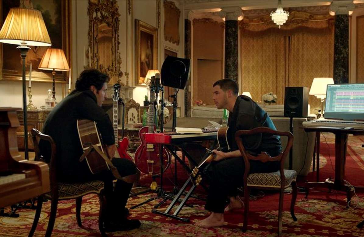 Muzičari, sede u salonskom stanu, drže instrumente, u pozadini se vidi računar sa jedene strane, a sa druge klavir.